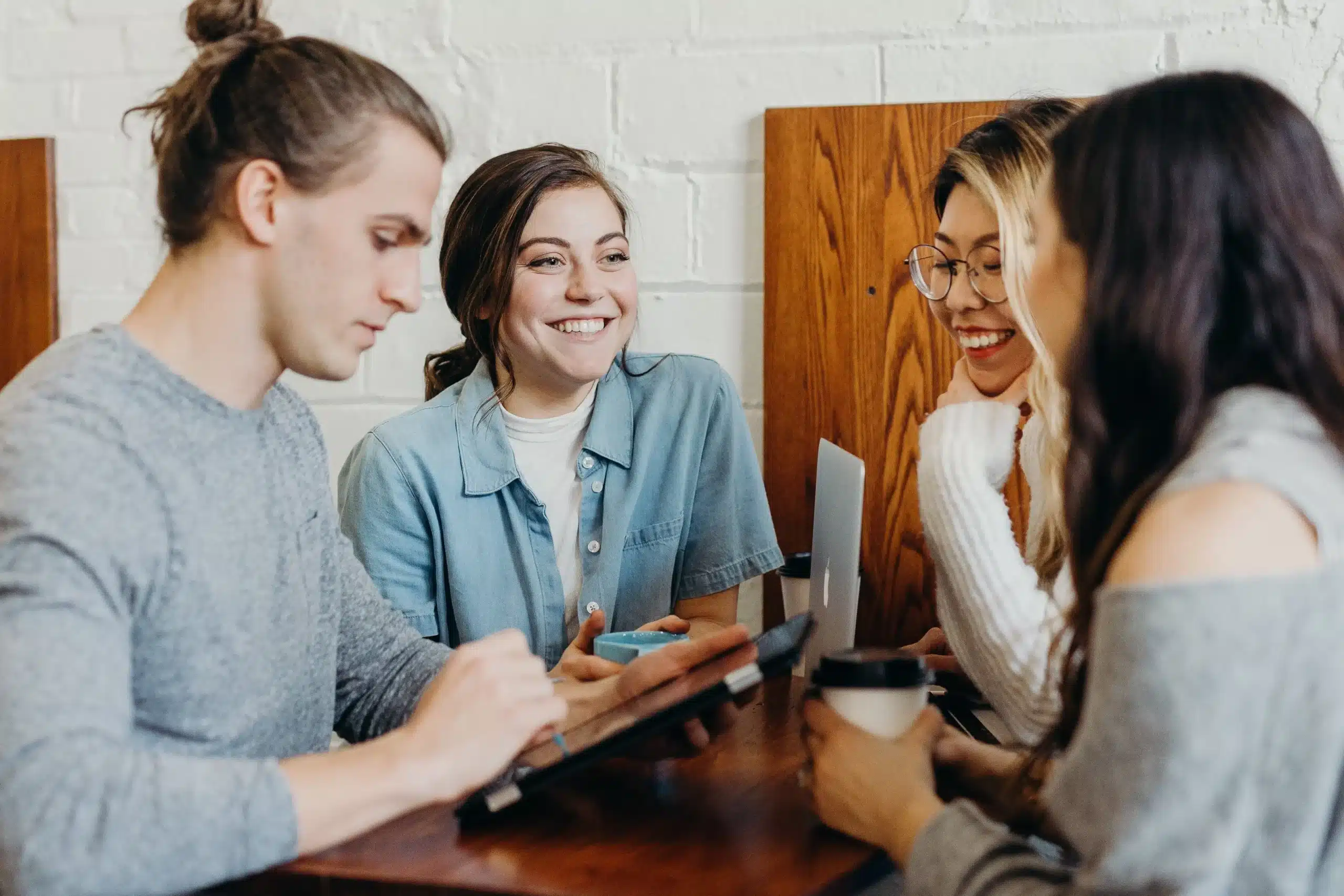 Een groep jonge mensen zit samen lachend aan tafel met laptops en koffie.