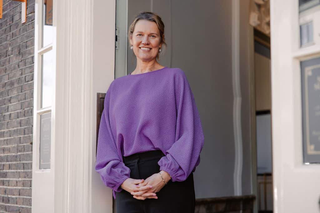 Vrouw in een paarse trui staat glimlachend in een deuropening.
