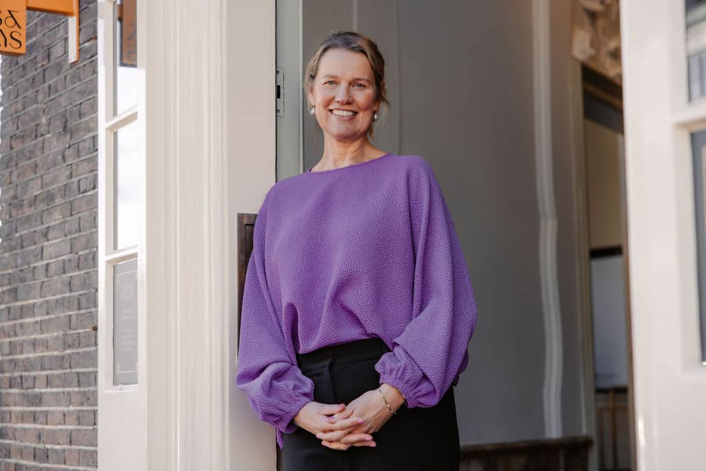 Een vrouw in een paarse trui staat glimlachend bij een deur.