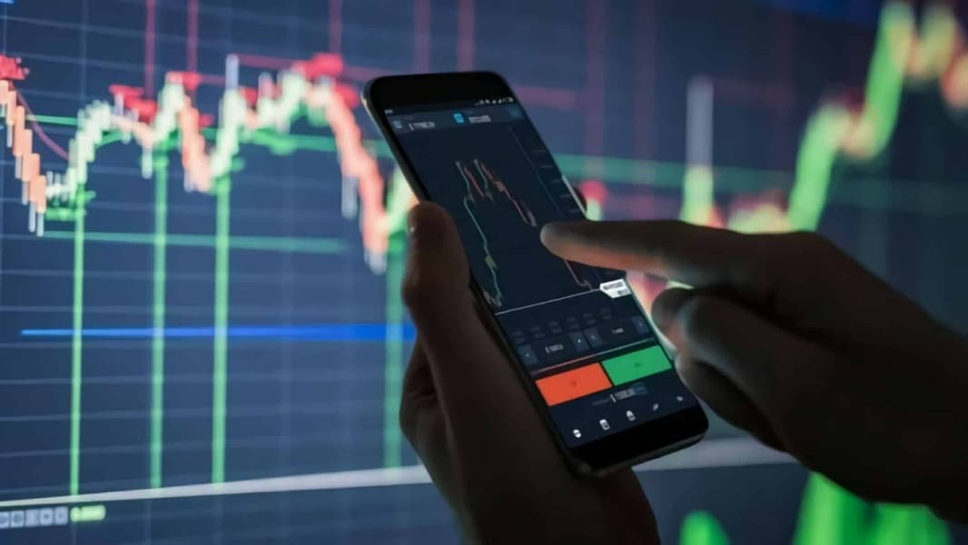 Een hand bedient een smartphone met daarop een handelsapplicatie, tegen de achtergrond van grafieken op een scherm.