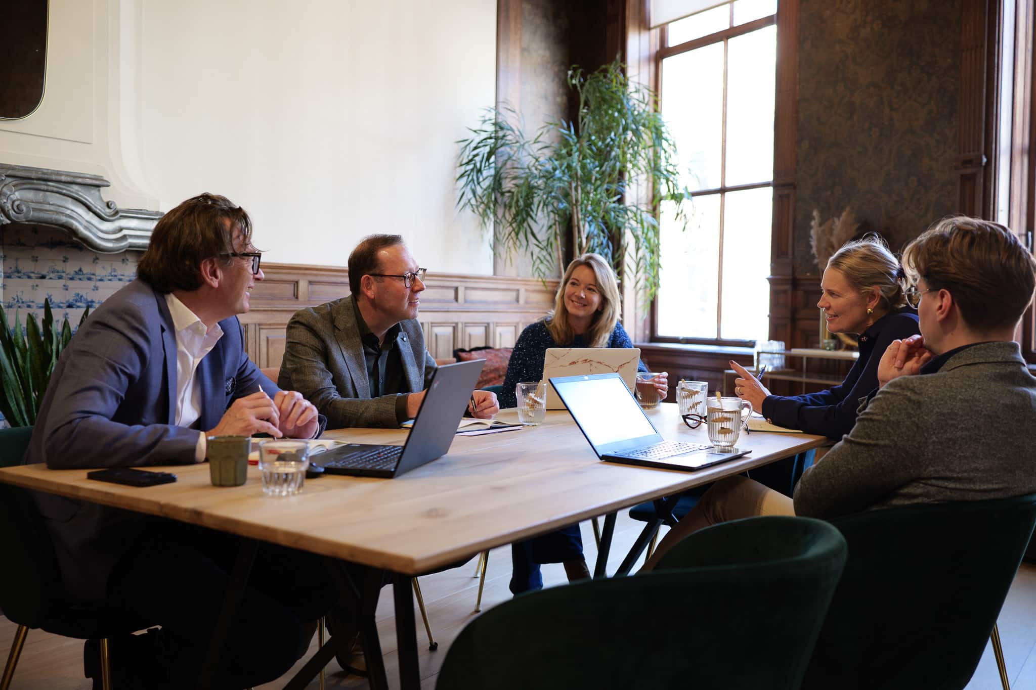 Een groep mensen vergadert aan een tafel in een stijlvolle kamer met laptops en notitieboeken.