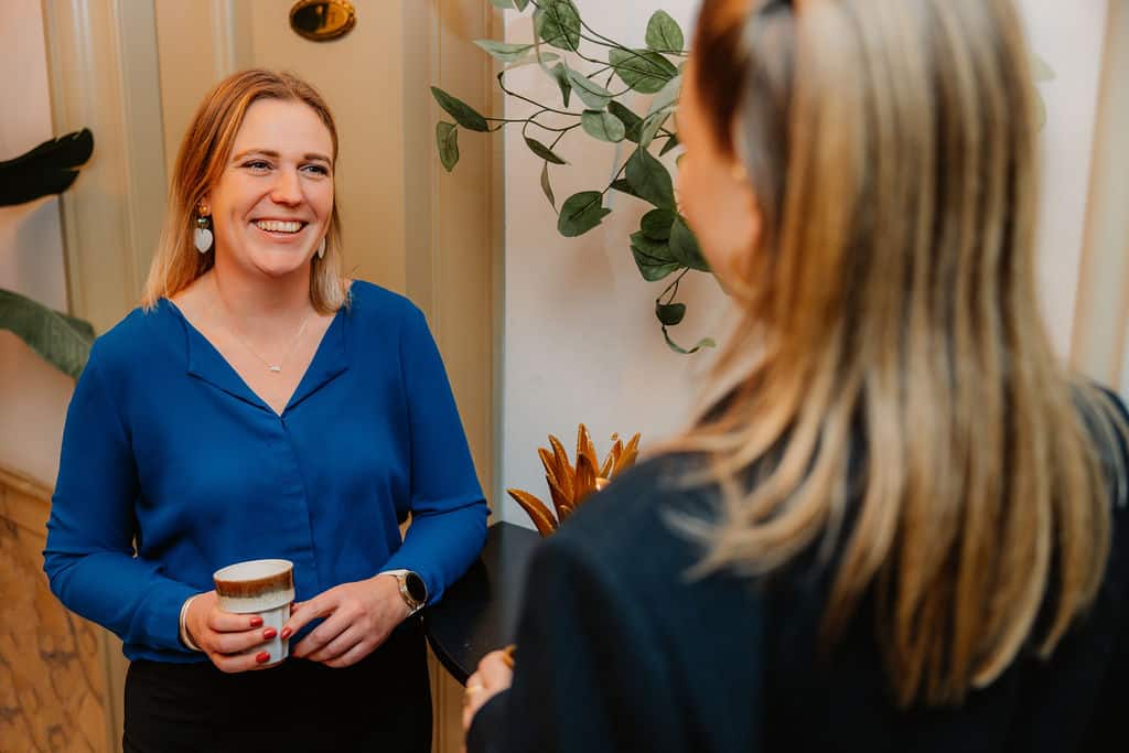 Vrouw in blauw overhemd lacht en houdt een kop vast tijdens een professioneel gesprek