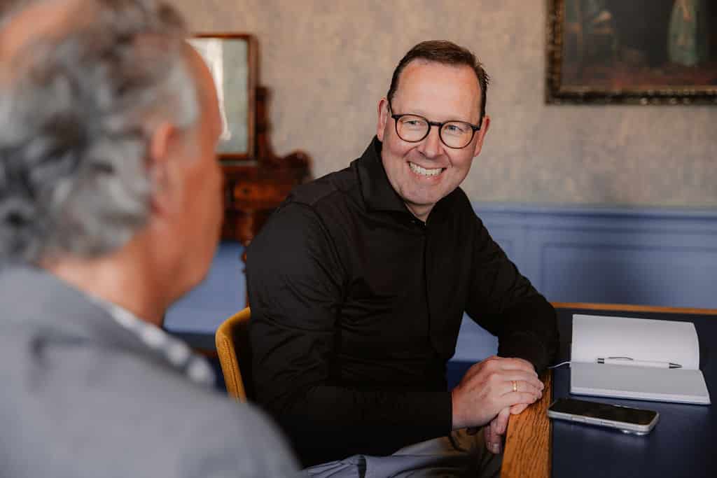 man in zwart overhemd die lacht naar zijn collega, leunend tegen een bureau met een opengeklapt schrift en mobiel.