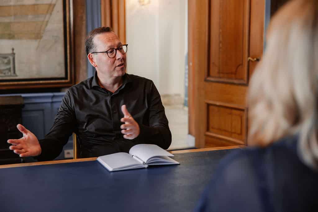 Man in donker overhemd en bril voert een gesprek aan een vergadertafel in een klassiek kantoor, met een opengeslagen notitieboek voor hem