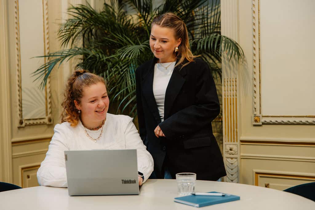 Twee vrouwen bekijken samen een laptop aan een vergadertafel in een klassiek kantoor