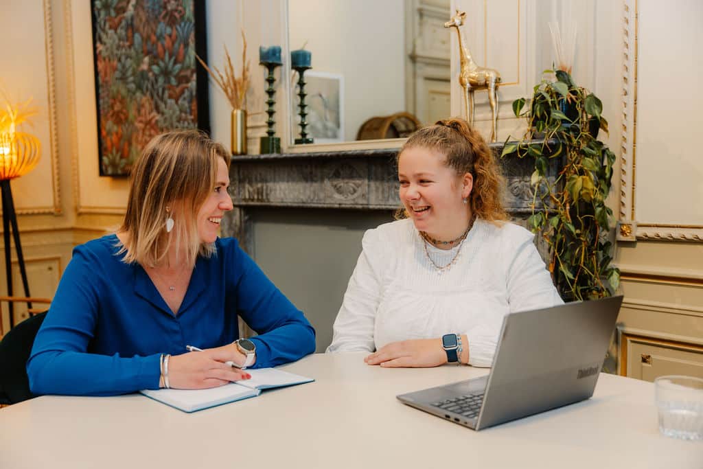 Twee vrouwen lachen samen aan een vergadertafel met een laptop in een klassiek kantoor met open haard
