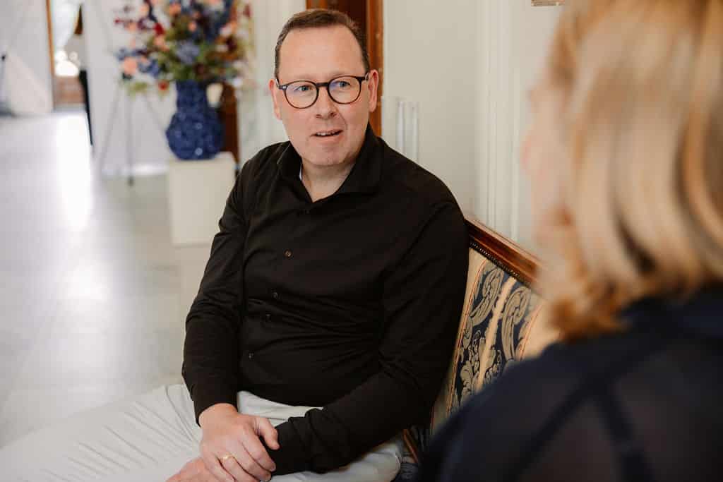 Man in donker overhemd en bril luistert aandachtig tijdens een gesprek in een klassiek interieur